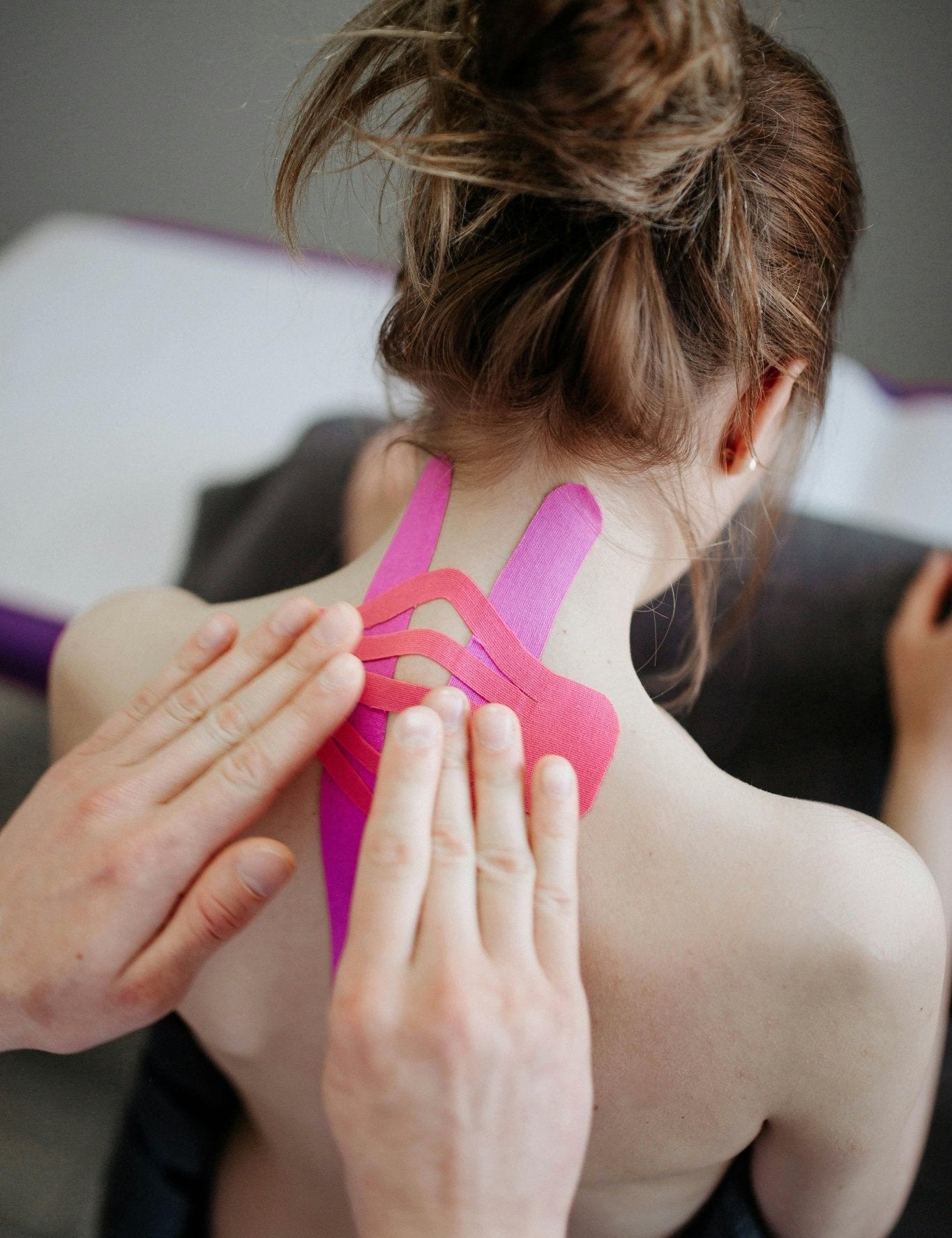 Physiotherapeut bringt Kinesio-Tape in Pink auf den oberen Rücken einer Frau auf – Methode zur Unterstützung für Narben und Beweglichkeit