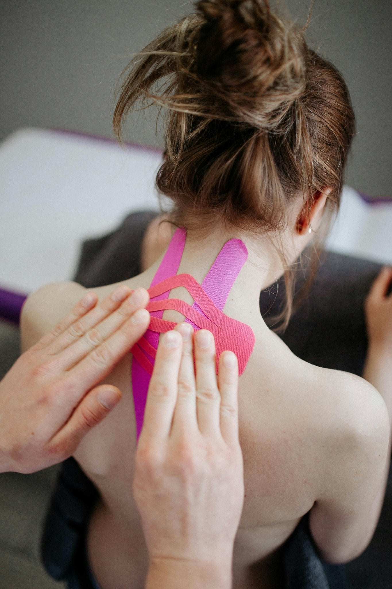 Physiotherapeut bringt Kinesio-Tape in Pink auf den oberen Rücken einer Frau auf – Methode zur Unterstützung für Narben und Beweglichkeit